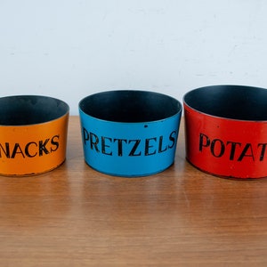 Peut inclure: Trois récipients métalliques ronds avec des inscriptions noires sur le côté. Les récipients sont orange, bleu et rouge. Les récipients sont étiquetés "Snacks", "Pretzels" et "Potato".