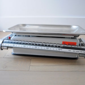 May include: A silver kitchen scale with a stainless steel weighing platform. The scale has a black dial with markings for grams and kilograms. The scale reads "bisieria" and "NOT LEGAL FOR TRADE".