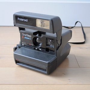 Peut inclure: Un appareil photo Polaroid OneStep Close-up noir avec un flash et une sangle. L'appareil photo est posé sur un sol en bois clair.