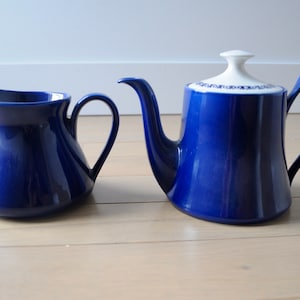 Pode incluir: Um conjunto de bule e jarra de creme de cerâmica azul. O bule tem uma tampa branca com um design floral azul. A jarra de creme tem uma forma simples e arredondada.