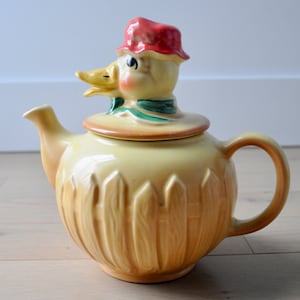 Może przedstawiać: Żółty ceramiczny czajnik z pokrywką w kształcie głowy kaczki. Kaczka ma czerwony kapelusz i zielone liście wokół szyi. Czajnik ma wzór płotu.