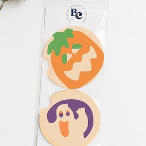 May include: Two circular car coasters in a clear plastic sleeve. The top coaster features an orange pumpkin with a green stem and a wavy mouth. The bottom coaster displays a purple ghost with an orange mouth. The brand logo Backroad Creative Co. Est. 2016 is printed on the white backing card.