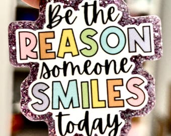 Puede incluir: Un pin morado brillante con una forma inusual, con el texto "Be the REASON someone SMILES today" en letras coloridas y negras. El pin está sostenido por una mano.