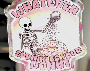 Puede incluir: Una pegatina transparente con un esqueleto espolvoreando chispas sobre una rosquilla rosa. El texto "WHATEVER SPRINKLES YOUR DONUT" está en letras rosas con burbujas. El fondo tiene un diseño de arcoíris.