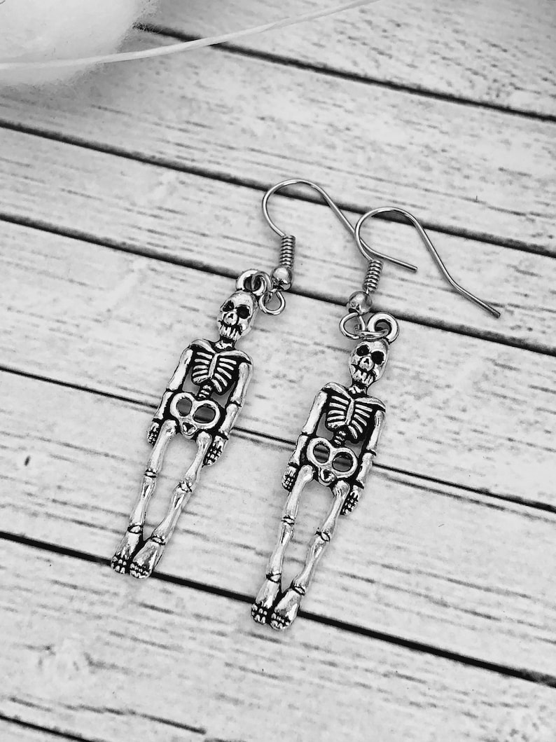 Puede incluir: Un par de pendientes de esqueleto de plata. Cada pendiente presenta un dise&ntilde;o de esqueleto detallado con una calavera en la parte superior.