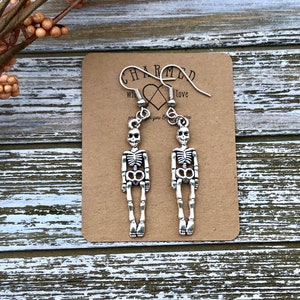 Puede incluir: Pendientes de esqueleto de plata colgando en una tarjeta marr&oacute;n con el texto "CHARMED with love".