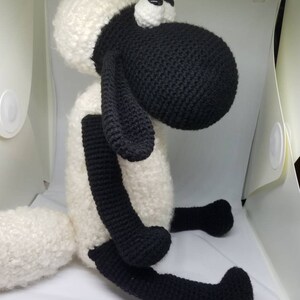 Puede incluir: Un juguete de peluche de oveja tejido a crochet con patas negras, cabeza negra y lana blanca. La oveja tiene grandes ojos negros y una cara blanca.