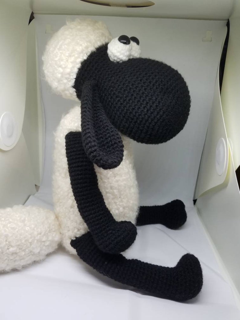 Puede incluir: Un juguete de peluche de oveja tejido a crochet con patas negras, cabeza negra y lana blanca. La oveja tiene grandes ojos negros y una cara blanca.
