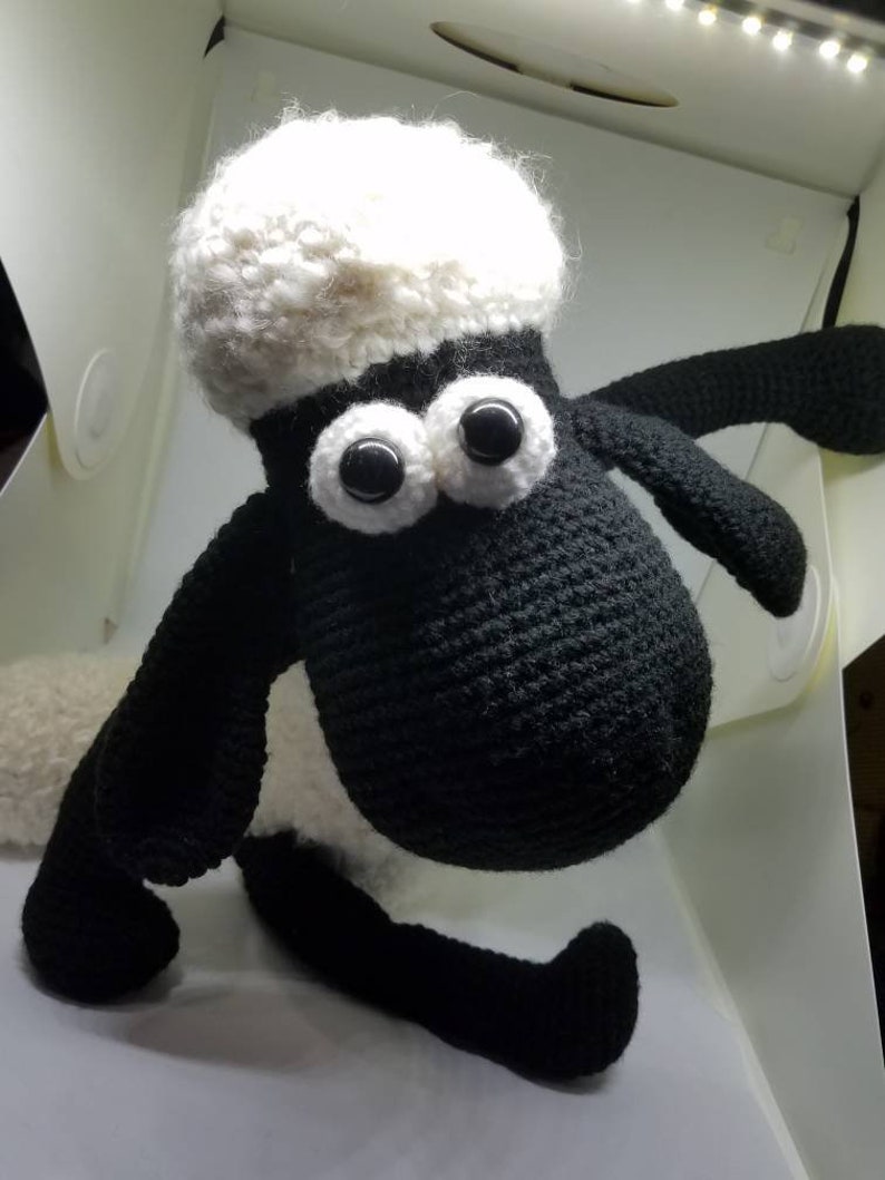 Puede incluir: Un juguete de peluche de oveja de crochet negro y blanco con grandes ojos negros. La oveja est&aacute; sentada sobre una superficie blanca.