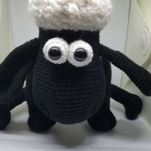 Puede incluir: Un juguete de crochet de oveja negro con lana blanca y grandes ojos negros. La oveja est&aacute; de pie sobre una superficie blanca.