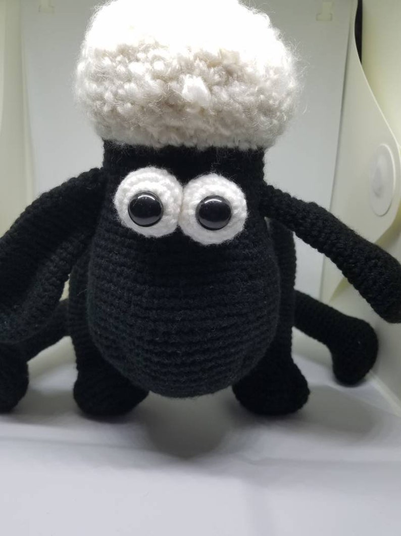 Puede incluir: Un juguete de crochet de oveja negro con lana blanca y grandes ojos negros. La oveja est&aacute; de pie sobre una superficie blanca.