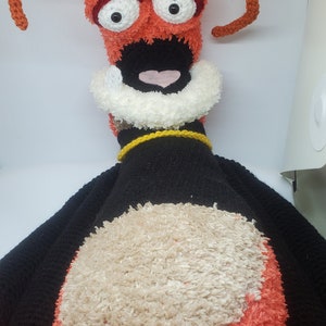 May include: A handmade plush toy with a whimsical design. The toy features a head with large white eyes, a black mouth, and orange fur. The body is black with a tan belly and orange feet. It has orange antennae and a white collar.