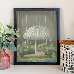 May include: Framed art print depicting a slug sheltering under a mushroom in a rainy forest. The illustration uses a muted colour palette of greens, browns, and greys. The artwork evokes a sense of calm and nature.
