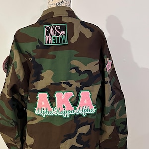 May include: A camouflage green utility jacket with a pink and green "AKA Alpha Kappa Alpha" patch on the back. The jacket also has a green patch that says "Oh So Pretty!" on the upper back.
