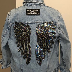 May include: Light blue denim jacket with sequined wing design on the back. The wings feature iridescent sequins with gold trim. A black patch with white text reads "I'M NOT PERFECT BUT I'M A LIMITED EDITION".