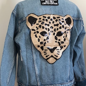 May include: A blue denim jacket with a gold and black sequined leopard head design on the back. The jacket has a patch on the back that says "I'm not perfect but I'm a limited edition."