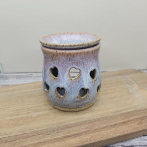 May include: A ceramic wax warmer featuring a speckled lavender and blue glaze with a glossy finish. The cylindrical body has cut-out heart shapes around the center and a removable dish on top, all with a rustic, earthy brown rim and base.