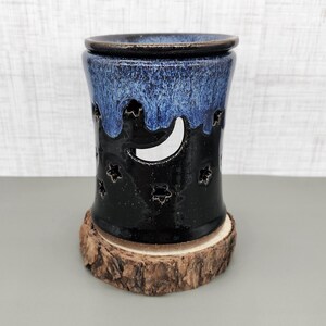 May include: A ceramic oil warmer with a dark blue and black glaze. The warmer features a crescent moon and star cutouts, and sits on a wooden base. The top is a darker blue, and the base is a natural wood slice.