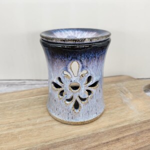 May include: A ceramic oil warmer with a dark blue and lavender glaze. The warmer has a decorative floral cutout design and a matching lid. The warmer is sitting on a wooden cutting board.