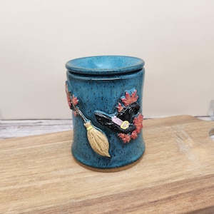 May include: A cylindrical ceramic wax warmer with a speckled teal blue glaze. The exterior features raised relief decorations including a tan broomstick, a black witch hat with a yellow buckle, and several small red maple leaves.