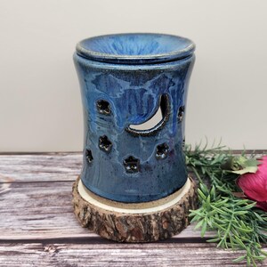 May include: A blue ceramic essential oil warmer with a crescent moon and star cutouts. The warmer sits on a wooden base. The ceramic has a glossy finish and is cylindrical in shape.