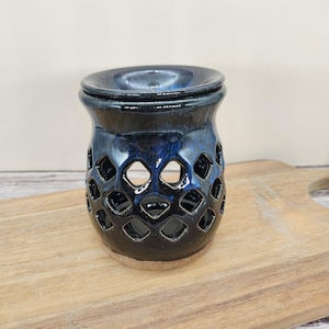 May include: A ceramic oil warmer with a dark blue glaze. The warmer has a round top and a body with diamond-shaped cutouts. The base is unglazed. The warmer sits on a wooden cutting board.