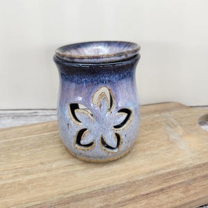 May include: A ceramic essential oil warmer with a flower-shaped cutout design. The warmer has a blue and purple glaze with a brown rim and base. The product is designed to hold essential oils and is approximately 4 inches tall.