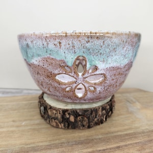 May include: A handmade ceramic bowl with a floral cutout design. The bowl features a speckled brown and turquoise glaze. It sits on a small, round wooden base, showcasing a rustic aesthetic. The bowl is displayed on a wooden cutting board.