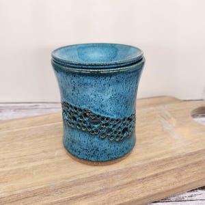 May include: A handcrafted, turquoise ceramic container with a lid. The cylindrical body features a textured, openwork band. The container sits on a wooden cutting board, showcasing its unique design and artisanal quality.