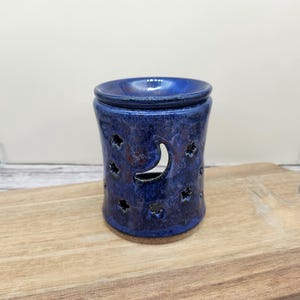 May include: A cylindrical ceramic wax warmer featuring a deep blue glaze with mottled dark undertones. The exterior is decorated with cut-out shapes of a crescent moon and multiple small stars, allowing light to pass through the hollow body.