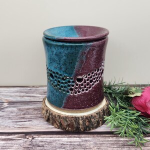 May include: A handcrafted ceramic candle warmer with a teal and burgundy color scheme. The cylindrical warmer features a perforated design and a matching lid. It sits on a rustic wooden base, perfect for home decor.