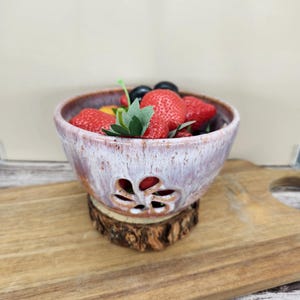May include: A ceramic fruit bowl with a floral cut-out design, filled with strawberries and other fruits. The bowl has a speckled, light purple and brown glaze. It sits on a wooden slice, placed on a wooden cutting board.