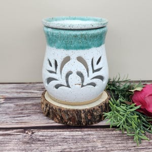 May include: A white and teal ceramic essential oil diffuser with leaf cutouts. The diffuser sits on a wooden slice and has a matching lid. The design is speckled with a teal rim. A decorative item for aromatherapy.