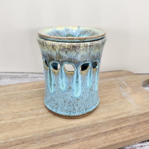 May include: A ceramic wax warmer with a blue and brown glaze. The cylindrical warmer has a series of oval cutouts around the middle and a lid. The warmer sits on a wooden cutting board.