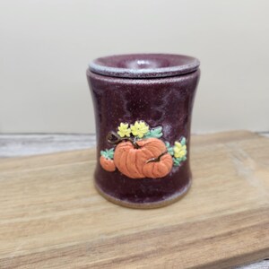 May include: A cylindrical ceramic wax warmer with a deep plum-colored glaze and a speckled finish. The front features a raised, three-dimensional orange pumpkin appliqué with small yellow flowers and green leaves attached to the side of the vessel.