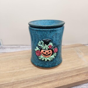 May include: A cylindrical ceramic wax warmer with a speckled teal blue glaze. The front features a raised, sculpted emblem of an orange jack-o'-lantern wearing a black top hat, surrounded by small green leaves and pink roses in relief.