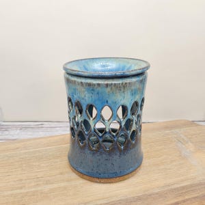 May include: A cylindrical ceramic wax warmer featuring a glossy blue glaze with brown speckles and a cutout diamond pattern around the center. The piece has a wide, shallow bowl at the top and a solid, unglazed tan base at the bottom.