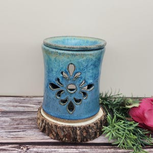 May include: A blue ceramic essential oil diffuser with a floral cutout design. The diffuser sits on a wooden slice base. The ceramic has a glossy finish and a lid. The background is a light color.