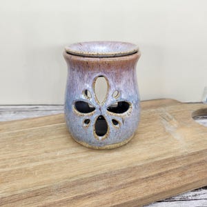 May include: A ceramic essential oil warmer with a floral cut-out design. The warmer has a two-tone glaze, with a brown top and a blue and purple body. The warmer sits on a wooden cutting board.