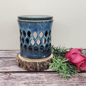 May include: A blue ceramic oil warmer with a honeycomb design, featuring a lid. The warmer sits on a wooden slice base. A red rose and greenery are in the background. The warmer is approximately 5 inches tall.