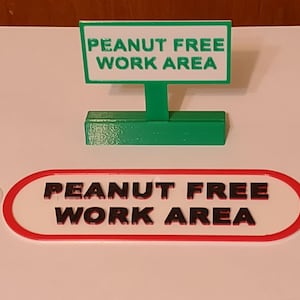 May include: A green and white sign that reads "PEANUT FREE WORK AREA" is displayed. Below it is a matching oval sign with the same text. A small suction cup hook is to the left of the oval sign.