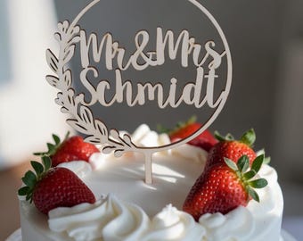 May include: A light-colored wooden cake topper featuring a circular frame with a leafy branch design on the left side. The text "Mr & Mrs Schmidt" is cut out in a cursive script font. The topper is inserted into a white frosted cake with strawberries.