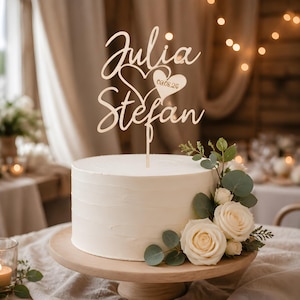 Könnte beinhalten: Ein runder weißer Kuchen auf einem Holzständer, gekrönt von einem lasergeschnittenen Holzkuchenaufsatz mit den Namen Julia und Stefan in Schreibschrift, einem Herz und dem Datum 09.08.26. Der Kuchen ist mit weißen Rosen und Eukalyptus verziert.
