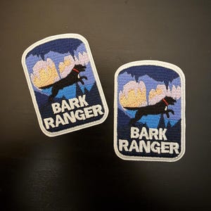 May include: Two embroidered patches with a navy blue background and a white border. The patches feature a black dog leaping over a mountain scene with the text "BARK RANGER" in white. The dog has a red collar.