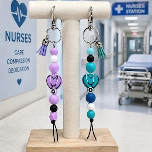 May include: Two beaded keychains with a medical theme. One keychain has purple, white, and black beads, a purple stethoscope charm, and a purple tassel. The other has teal, black, blue, and white beads, a teal stethoscope charm, and a teal tassel. The word "Nurses" is visible.