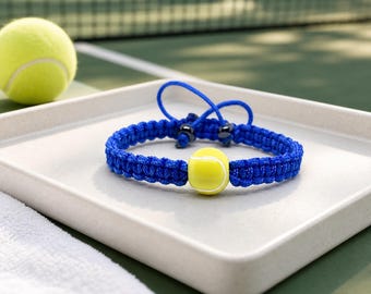 May include: A blue braided bracelet with a yellow tennis ball bead. The bracelet is displayed on a white tray, with a tennis ball and a tennis court in the background. The bracelet is a sporty accessory.