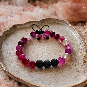 May include: A bracelet with dark pink, light pink, and black beads. The bracelet is presented on a light brown, textured plate, with a pink background. The beads are likely 0.8 cm in diameter.