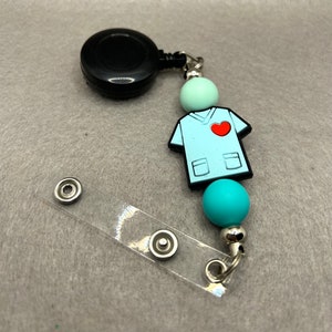 May include: A retractable badge reel with a black reel, a clear plastic badge holder, and a blue and green beaded chain with a blue scrub top charm with a red heart on the pocket.