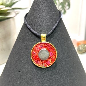 May include: A gold-toned pendant necklace with a red and pink design, featuring a silver-colored center. The pendant hangs from a black cord. The necklace is displayed on a black stand.