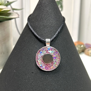 May include: A round pendant necklace with a black cord. The pendant has a silver-colored bezel filled with pink and iridescent glitter. A dark gray circle is in the center of the pendant.